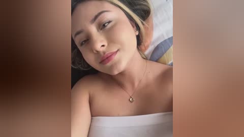 Media: A video of a young, light-skinned woman with brown hair lying on a bed, wearing a strapless white dress and a delicate necklace, with a blurred person's hand visible.