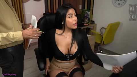 Media: Video of a Latina woman with long black hair, large breasts, wearing a black crop top, beige skirt, and thigh-high stockings, being held by a man in an office.