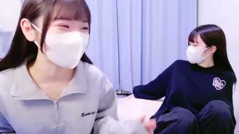 Media: Video of two young Asian women in white face masks, one in a light gray zip-up hoodie, the other in a black sweatshirt, sitting on a bed in a dimly lit room with blue curtains.