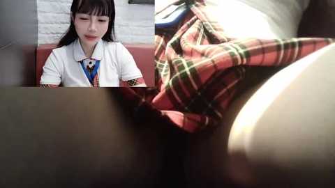 Media: A video of a young Asian woman with straight black hair, wearing a white shirt and a plaid skirt, seated on a red couch. The background features a white brick wall.