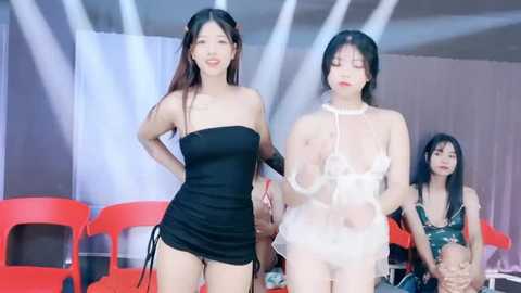 Media: Video of two Asian women in revealing black and white outfits, standing confidently in a brightly lit, modern indoor setting with red plastic chairs in the background.