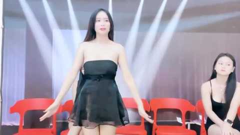 Media: Video of two Asian women in black dresses, one with long sleeves and a sheer skirt, the other in a sleeveless dress, on a stage with red chairs and white lights in the background.