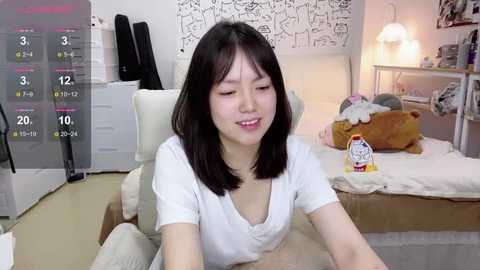 Media: Video of an East Asian woman with shoulder-length black hair, wearing a white t-shirt, sitting on a white chair in a cozy, cluttered bedroom with a teddy bear and a stuffed animal on the bed.