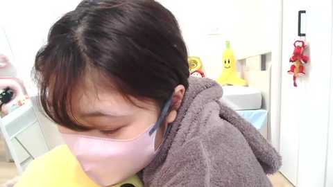 Video of an Asian woman with short, dark hair wearing a pink surgical mask and brown fuzzy robe, leaning over a yellow object in a brightly lit room with toys and a door.