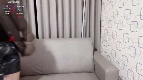 Media: Video of a beige sofa with a person in black leather attire, standing, in a modern room with geometric-patterned wallpaper.