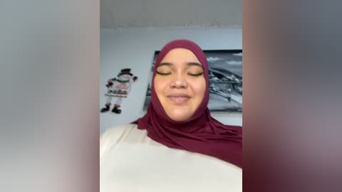 Media: A video of a smiling, light-skinned woman wearing a burgundy hijab and white top, standing indoors with a white wall and a black-and-white framed photo in the background.