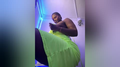 Media: Video of a dark-skinned woman with braided hair in a neon green dress, captured from a low angle, in a room with blue and purple lighting.