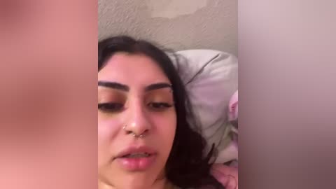 Media: Video of a young woman with long dark hair, light brown skin, and full lips, lying on a bed with pink and white pillows, nose ring visible.