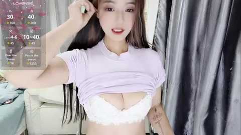 Media: Video of an East Asian woman with long black hair, fair skin, and a slim physique. She is lifting her white lace bra, revealing cleavage. Background features a digital clock, a white couch, and dark curtains.