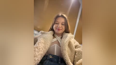 Video of a young woman with light skin, brown hair, and a soft smile, wearing a beige fur coat and high-waisted jeans, standing in a warmly lit room with a beige wall.