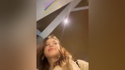 A video of a young woman with long, wavy brown hair and a light complexion, wearing a beige sweater, standing in a dimly lit room with a triangular skylight emitting a bright beam of light.