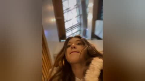 Media: A video of a young woman with long brown hair, wearing a white fluffy bathrobe, lying on a bed in a dimly lit room with a wooden door and a window.