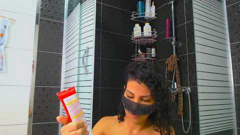 Media: Video of a young woman with curly black hair, wearing a face mask, holding a red tube of cream in a modern bathroom with black tiles and a shower with shelves.