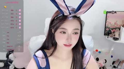 Media: A video of an East Asian woman with fair skin, long black hair, and bunny ears headband, wearing a light pink and blue bunny-themed outfit, seated indoors with a cluttered desk and calendar in the background.