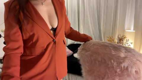 Media: A video of a woman in a rust-colored blazer, partially revealing a black bra, petting a fluffy pink dog, against white curtains and a lit lamp.