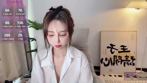 Media: A video of an East Asian woman with fair skin, red lipstick, and brown hair in a messy bun. She's in a white shirt, with a background featuring a potted plant, a framed painting, and a dimly lit room.