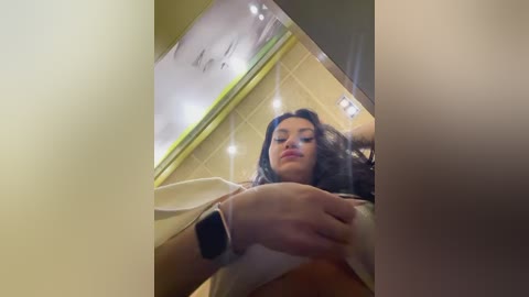 Media: Video of a woman with long dark hair, wearing a white top and a smartwatch, captured from a low-angle perspective, inside a bright, modern room with a white ceiling and recessed lighting.