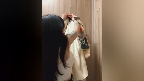 Media: Video of a woman with long, straight black hair, wearing a white sleeveless top, hanging a beige and black scarf with \"YOGA\" written on it on a wooden door.