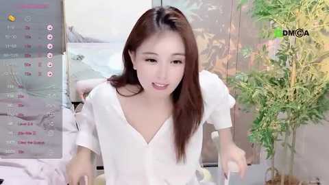 Media: Video of an East Asian woman with long brown hair, wearing a white blouse, smiling, seated in a living room with a plant and a TV screen showing a weather forecast in the background.