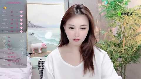 Media: Video of an Asian woman with long brown hair, wearing a white shirt, indoors with a potted plant and a TV screen showing a beach scene.