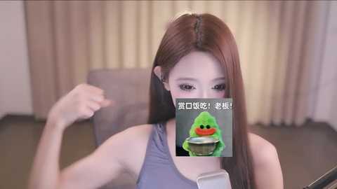 Media: Video of a young Asian woman with long brown hair, wearing a sleeveless grey top, covering her mouth with a green plastic toy frog. Background features beige curtains and a grey chair.