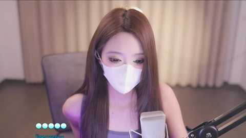 Media: Video of an East Asian woman with long, straight brown hair wearing a face mask, seated at a desk with a microphone, looking down at a laptop screen.