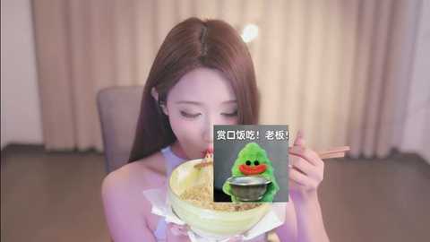 Media: Video of an Asian woman with long brown hair, wearing a white dress, eating from a bowl with chopsticks. The background features beige curtains and a chair. A meme with a green frog is overlaid.