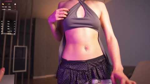 Media: Video of a slender, light-skinned woman with a navel piercing, wearing a grey halter top and black polka-dot skirt, standing in a dimly lit room.