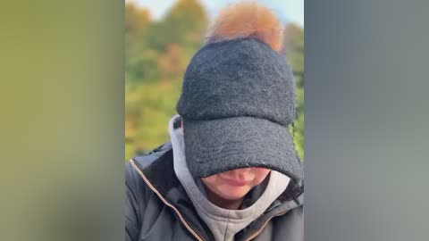 Media: A close-up video of a person wearing a gray knitted beanie with a pom-pom, a gray jacket, and a white scarf, with a blurred background of greenery and trees.