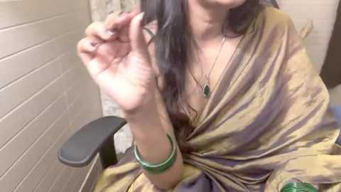 Media: A video of a woman with long, dark hair, wearing a gold sari and green bangles, brushing her hair, seated on a black chair in a tiled room.