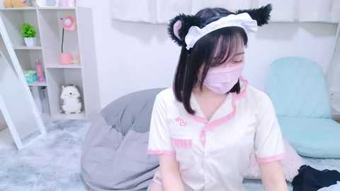 Media: Video of an East Asian woman in a white nurse uniform, cat ears, and mask, seated on a gray beanbag chair, in a softly lit, pastel-colored bedroom with shelves and plush toys.