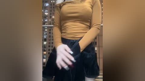 Media: A video of a woman in a mustard-yellow long-sleeve top and black skirt, holding a black purse, standing indoors with a window showcasing a cityscape at night.