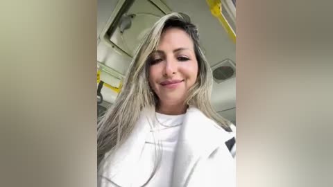 Media: Video of a smiling, fair-skinned woman with long, straight, blonde hair, wearing a white lab coat. Background shows a sterile, white-lit medical or scientific setting with yellow safety equipment.