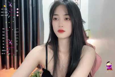 Media: Video of an East Asian woman with long black hair, fair skin, and red lipstick, wearing a black spaghetti strap top, sitting in a modern room with dark curtains.