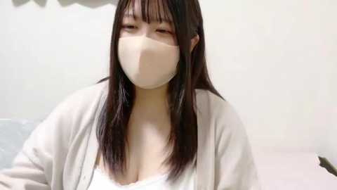 Media: Video of a young East Asian woman with long, straight black hair and fair skin, wearing a white mask, white cardigan, and a low-cut white top, sitting indoors on a bed with white sheets.