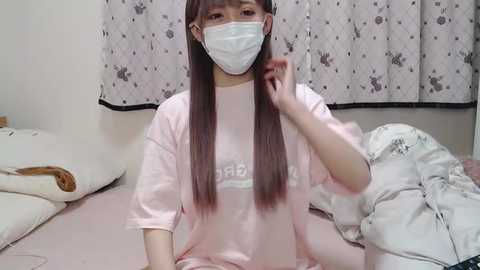 Media: Video of a young Asian woman with long, straight brown hair, wearing a white mask, pink T-shirt, and sitting on a bed with white sheets and pillows.