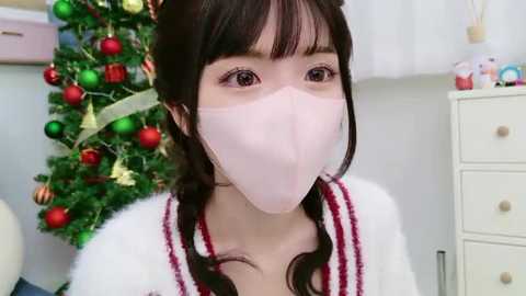 Media: A young Asian woman with long black hair in braids wears a pink surgical mask, a white robe with red trim, and stands in front of a decorated Christmas tree with red and gold ornaments.