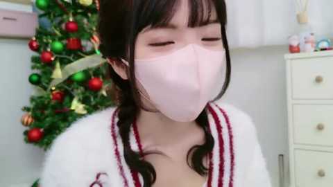 Media: A video of an Asian woman with long black hair, wearing a pink surgical mask, white robe, and a Christmas tree in the background.