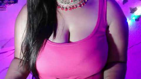 Media: Video of a curvy woman with medium brown skin, wearing a tight pink tank top that accentuates her large breasts and a beaded necklace, against a purple-lit background.