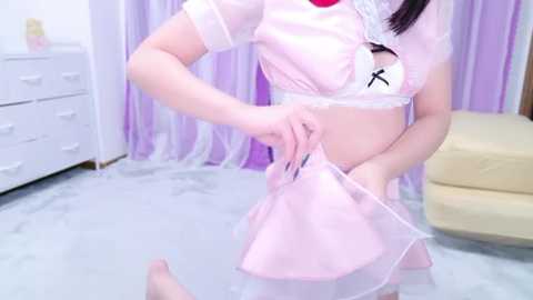 Media: Video of an Asian woman in a pink maid cosplay outfit, revealing a white bra with black bow, adjusting her skirt in a bright, pastel-colored room.