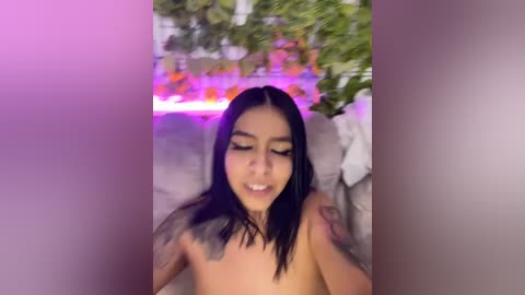 Media: Video of a young Asian woman with long black hair, wearing minimal makeup, smiling, and sitting outdoors in a garden with purple lighting.