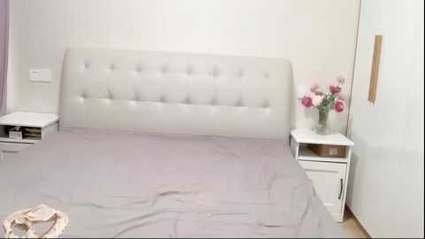 Media: Video of a minimalist bedroom with a tufted, white, upholstered headboard, grey bedding, white nightstands, and a vase of pink flowers.