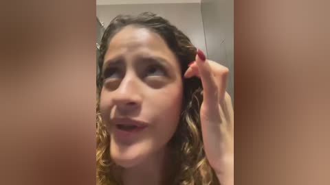 Media: Video of a young woman with curly brown hair, light skin, and red nails, looking distressed, holding her ear, in a blurry bathroom setting.