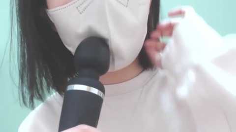 Media: Video of an Asian woman with long black hair, wearing a white mask and a white top, using a black vibrator, blurred background.