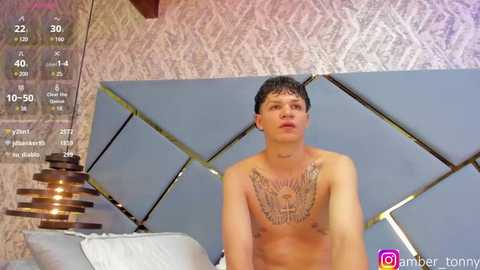 Media: Video of a shirtless, young, Caucasian man with short, dark hair, wearing a tribal tattoo on his chest. He stands in front of a mirrored, geometric headboard in a modern bedroom.