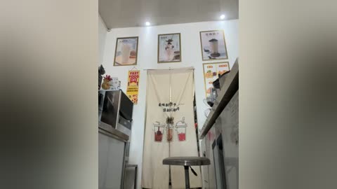 Media: Video of a narrow, dimly lit kitchen with white walls, framed posters, and a beige curtain with Thai writing. Stainless steel appliances and a stool occupy the space.