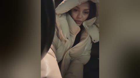 Media: Video of an Asian woman with a serious expression, wearing a puffy white coat and black pants, partially obscured by a blurred figure in the foreground.