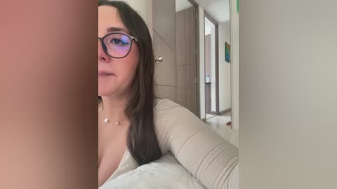 Media: A video of a woman with long brown hair, wearing glasses and a beige top, lying on a bed in a modern bedroom with light-colored walls and wooden doors.