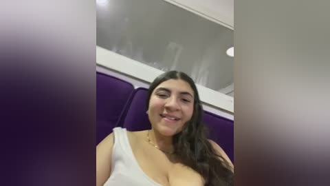 Media: Video of a young woman with long dark hair, smiling, wearing a white tank top, sitting on a purple sofa in a modern interior.