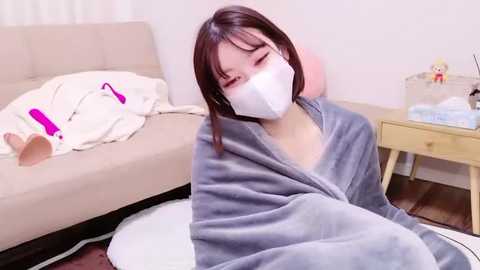 Media: Video of an Asian woman with straight, shoulder-length brown hair, wearing a white mask, gray robe, and sitting on a wooden floor beside a beige sofa and a small wooden nightstand with a stuffed animal.